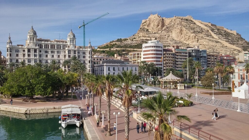 Scenic view of Alicante Marina with Santa Barbara Castle, historic buildings, and a carousel by the sea
